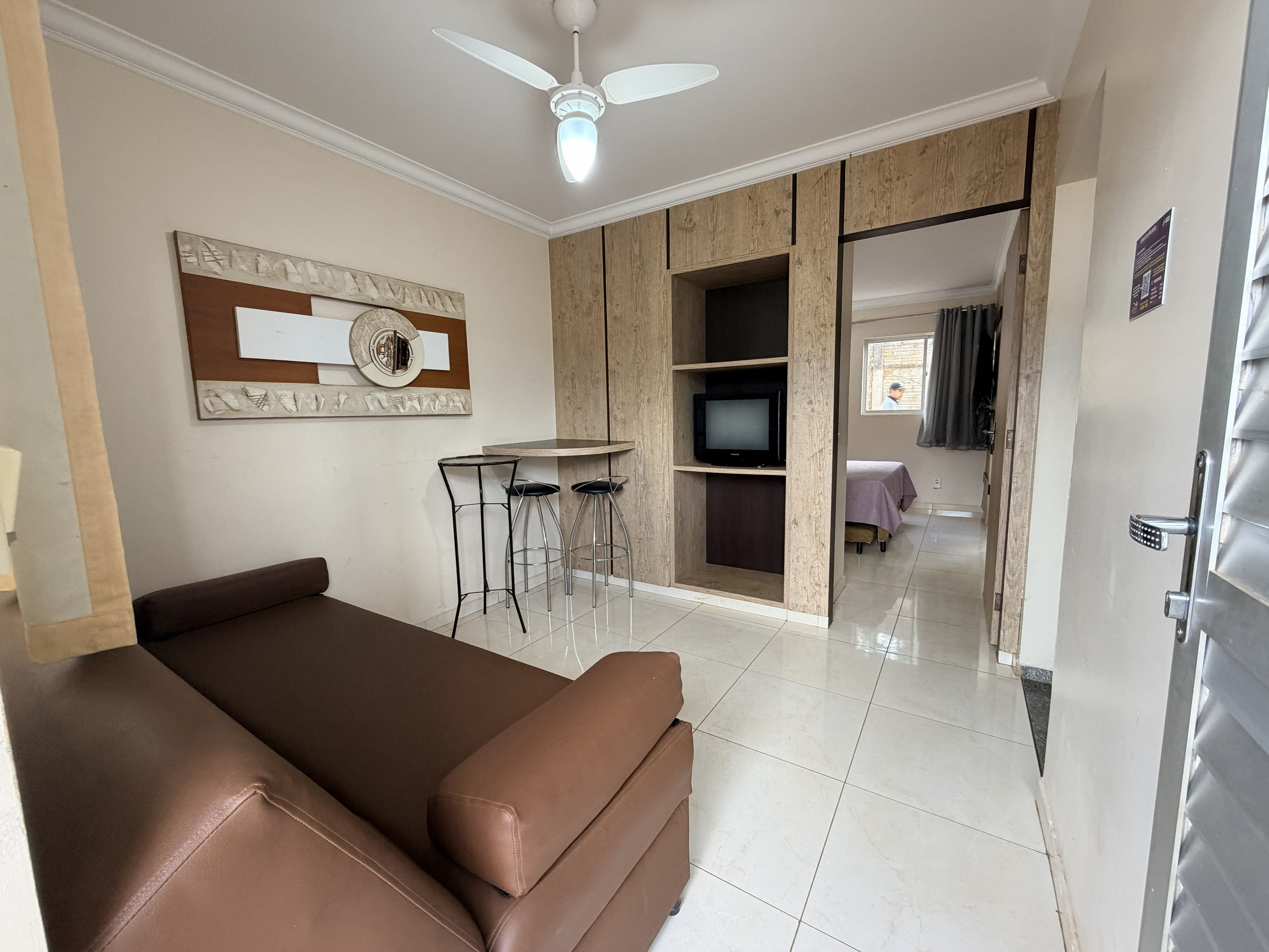 A modern living room with a brown sofa, a ceiling fan, a TV on a wooden shelf, and a dining area with bar stools.