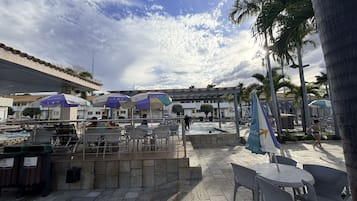 Uma área à beira da piscina com mobiliário e guarda-sóis, cercada por palmeiras e edifícios.