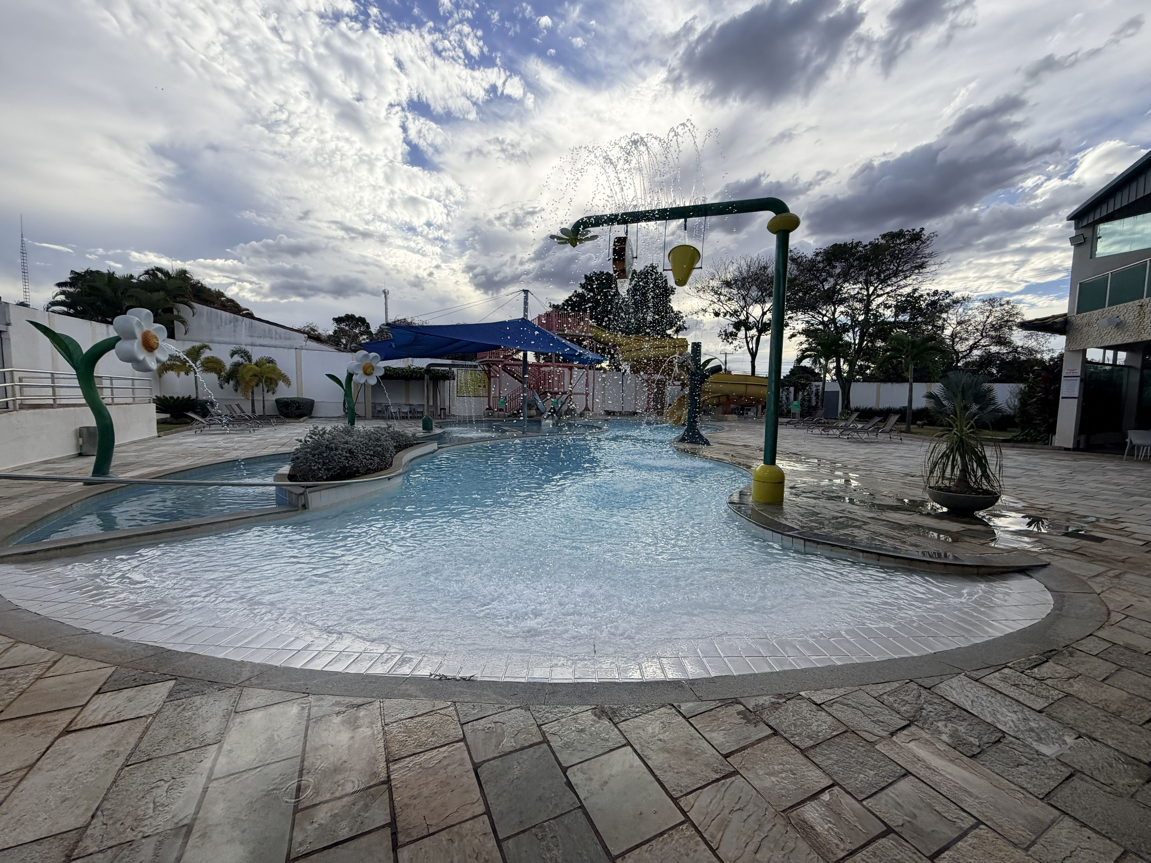 A water feature with a fountain and flower-shaped sculptures, surrounded by a paved area with seating and a building in the background.
