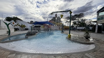 A water feature with a fountain and flower-shaped sculptures, surrounded by a paved area with seating and a building in the background.