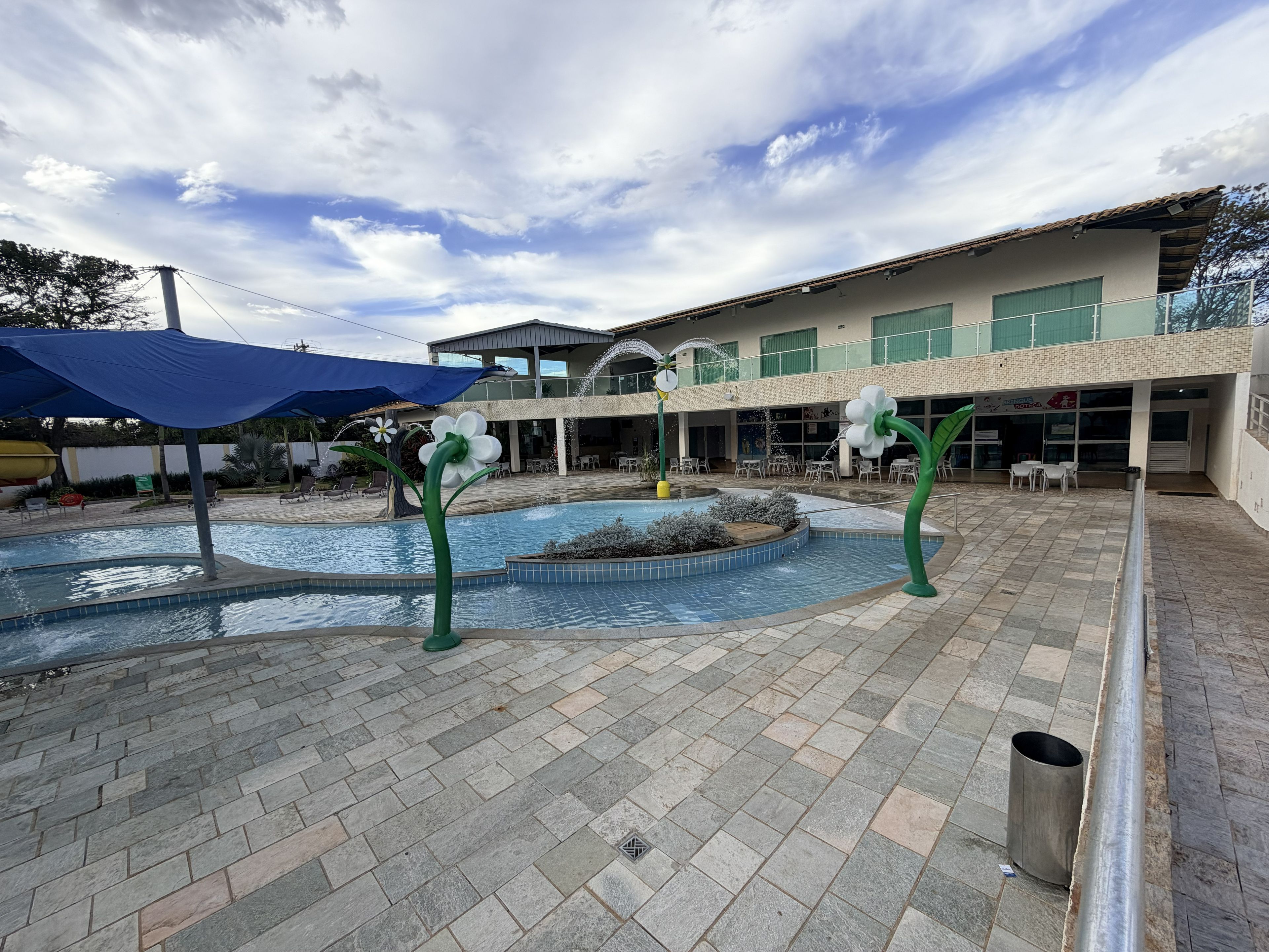 A swimming pool with a fountain feature, surrounded by a paved area with chairs and umbrellas.
