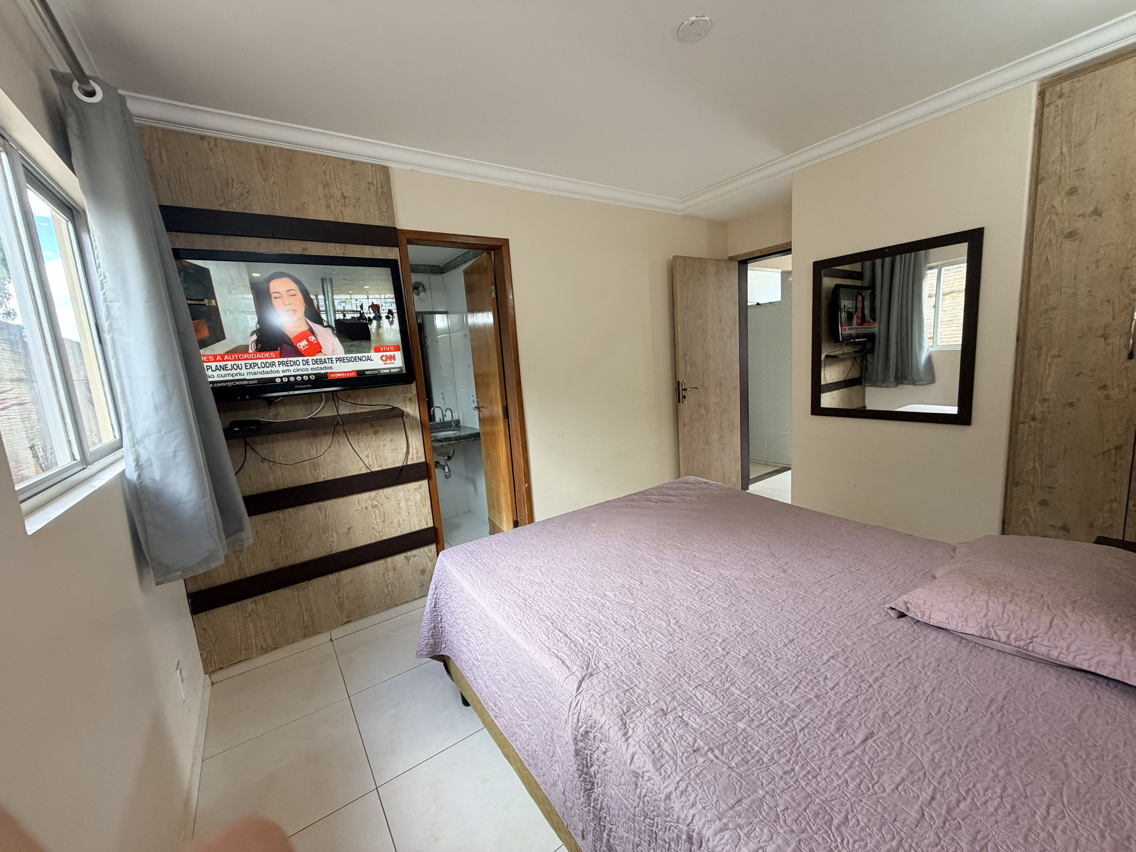 Una camera da letto con un letto, una televisione fissata a un pannello di legno, una finestra con tende e una porta che dà su un'altra stanza.