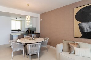Ein modernes Wohnzimmer mit einem runden Esstisch, weißen Korbstühlen, einer Couch mit Kissen, einer Küchenzeile mit Mikrowelle und einem großen abstrakten Gemälde an der Wand.