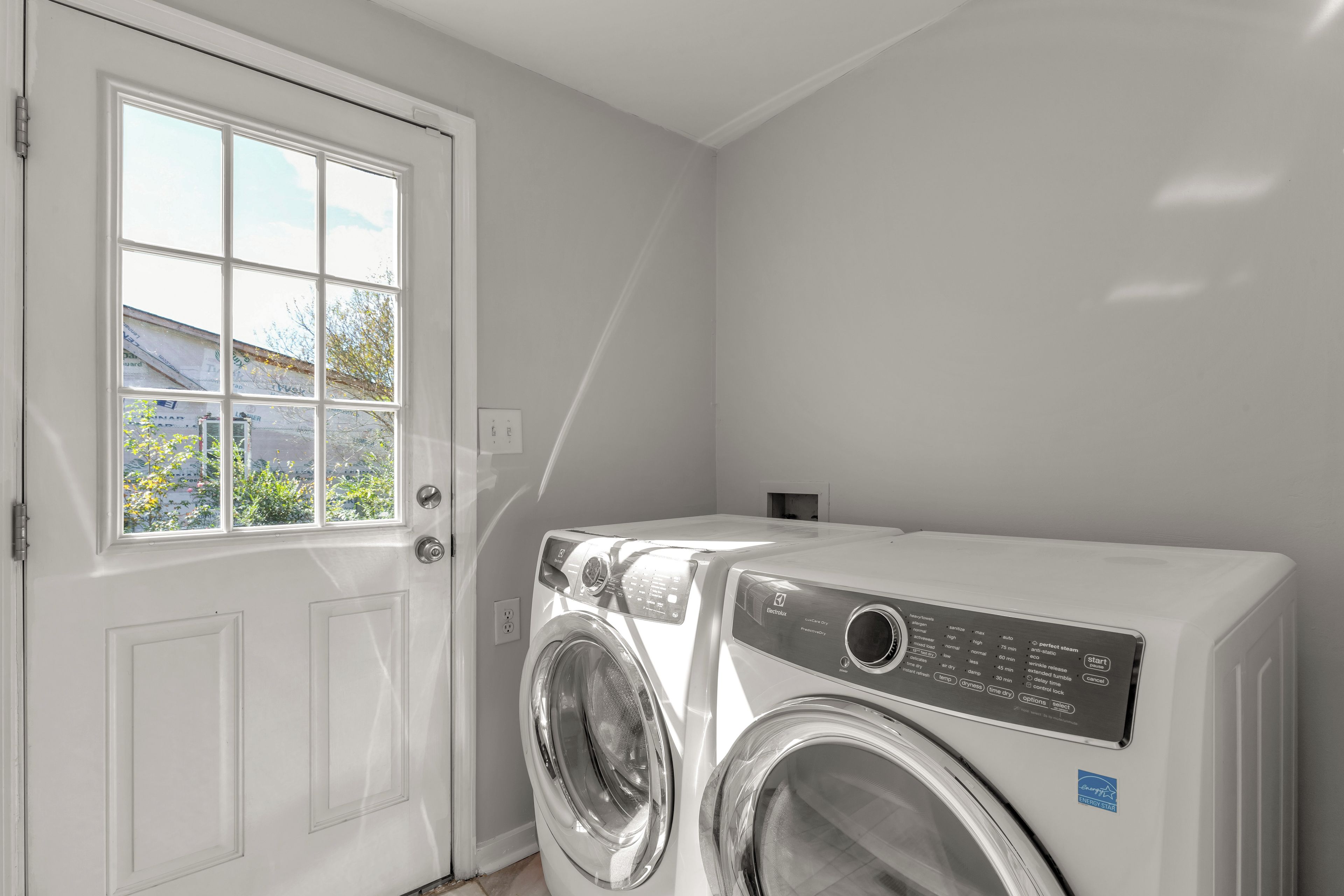 A laundry room with a front-loading washing machine, a door with a window, and a wall-mounted electrical outlet.