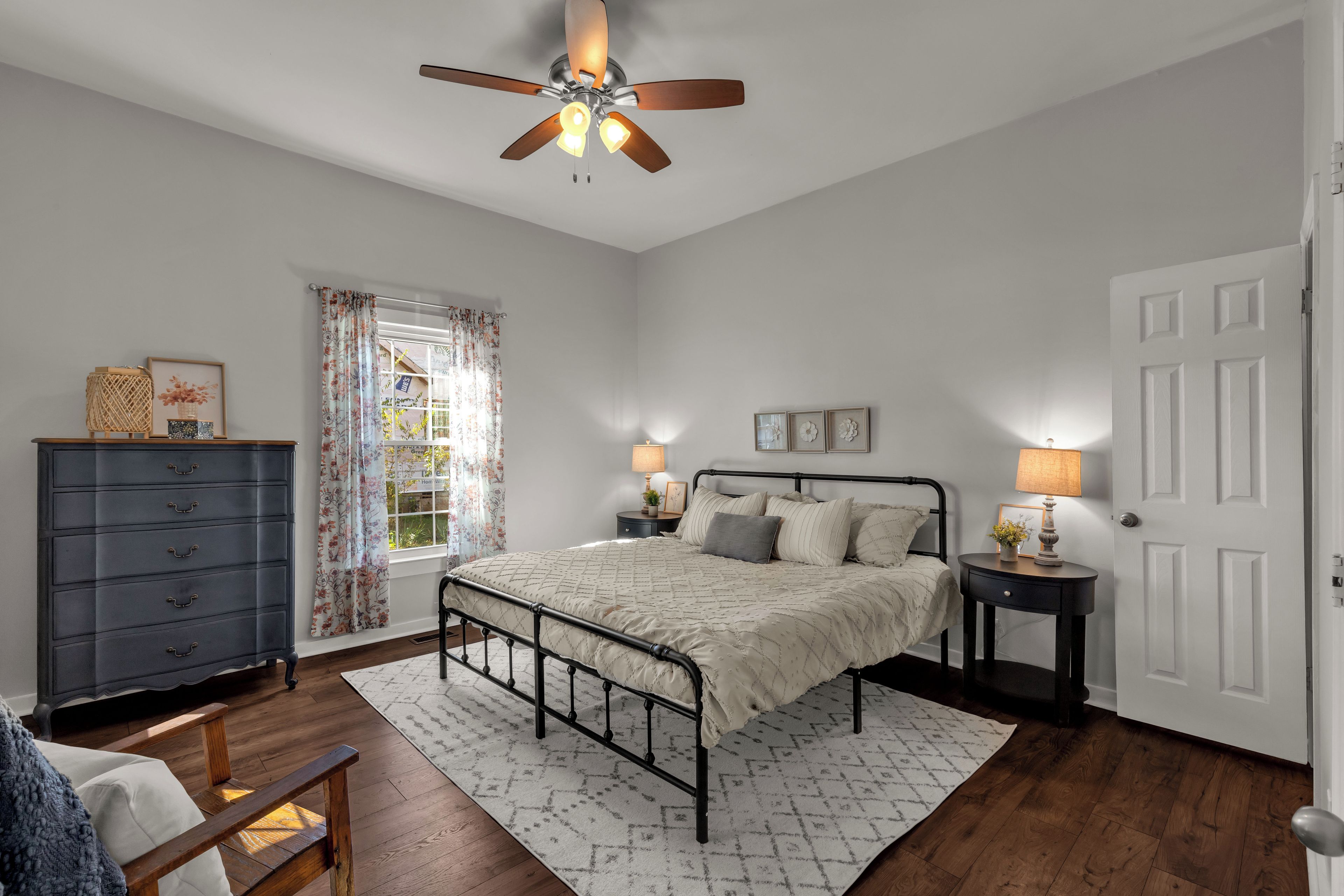 A bedroom with a bed, a dresser, a window with curtains, a ceiling fan, and a door.