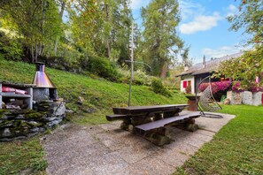 En utomhusplats med sittplatser, bestående av en bänk i sten och ett picknickbord, omgiven av grönska och en liten byggnad med en skorsten.