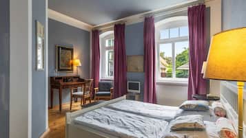 Een slaapkamer met een bed, een bureau en een stoel. Er is een raam met gordijnen, een lamp en kunst aan de muur.