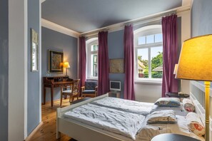 A bedroom with a bed, a desk, and a chair. There is a window with curtains, a lamp, and artwork on the wall.