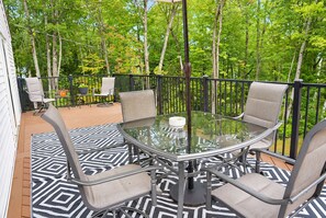 An outdoor patio with a glass table surrounded by chairs, a patterned rug, and a view of a wooded area.