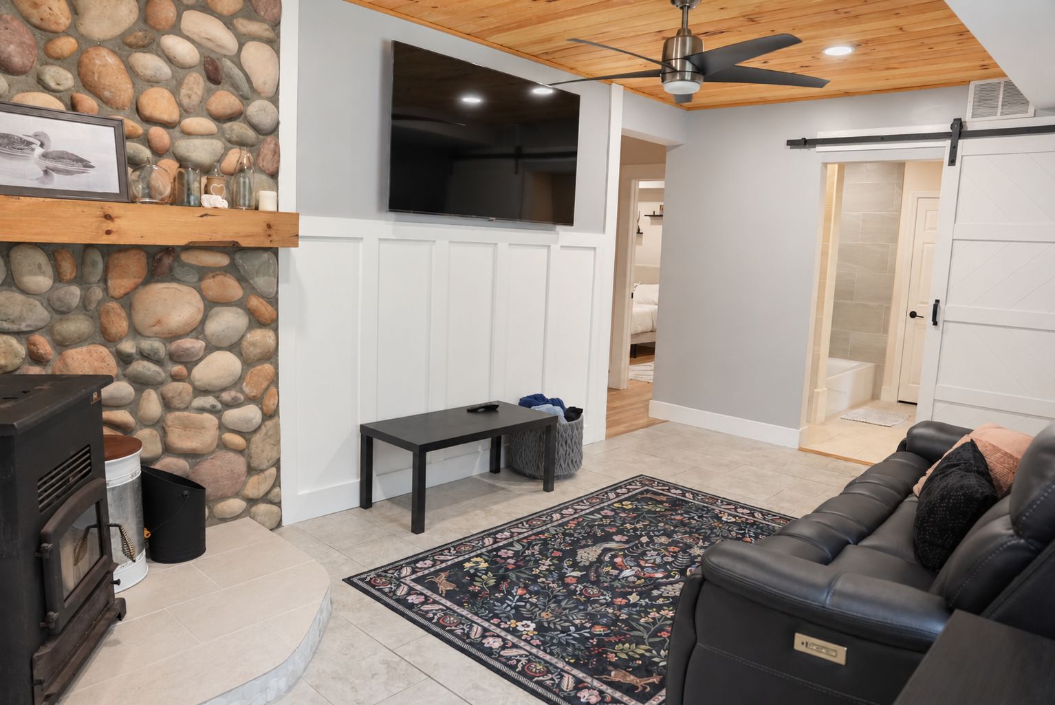 A living room with a stone wall, a flat-screen TV, a ceiling fan, a fireplace, a sofa, and a patterned rug.