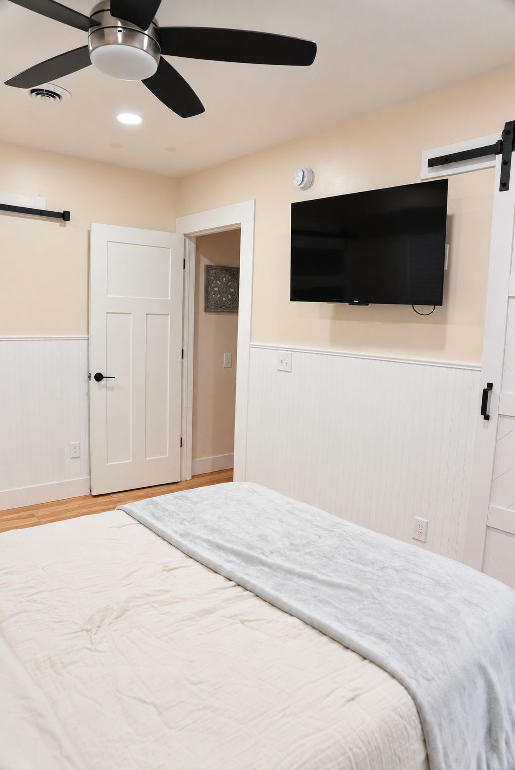 A bedroom with a bed, a ceiling fan, a television mounted on the wall, and a door leading to another room.