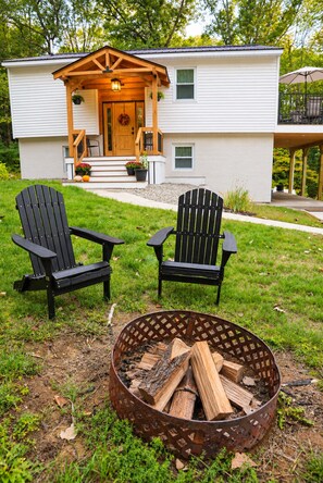 Une maison avec une véranda, deux fauteuils Adirondack et un foyer extérieur avec du bois.