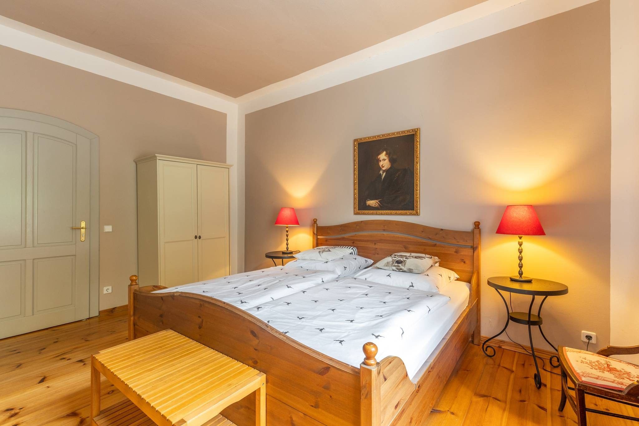 Une chambre à coucher avec un lit en bois, deux tables de chevet avec des lampes, un banc, une armoire et un tableau encadré au mur.