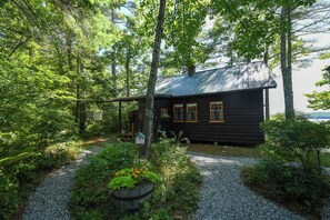 A cabin surrounded by dense forest, with a gravel path leading to the entrance. - NEW FOR 2027: Secluded Lakefront Cabin w/ Incredible Beach on Pleasant Lake (Otisfield)