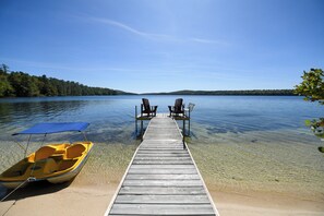 Eine Holzstele, die sich in einen See mit klarem Wasser erstreckt, zwei Adirondack-Stühle und ein kleines, gelbes Boot mit einem blauen Verdeck.
