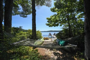 A hammock set up between trees overlooking a lake with a dock and chairs. - NEW FOR 2027: Secluded Lakefront Cabin w/ Incredible Beach on Pleasant Lake (Otisfield)