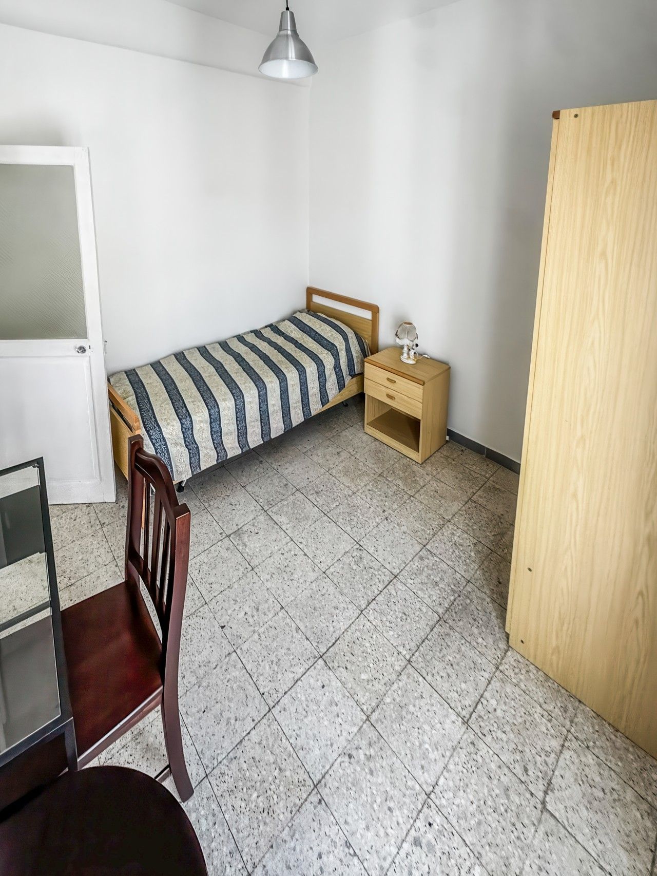 A small, sparsely furnished room with a bed, a nightstand, a chair, and a door.