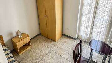 A room with a wooden wardrobe, a small wooden table with a chair, a sofa, and a hanging light fixture.