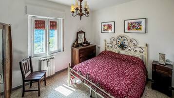 A bedroom with a bed, a chair, a dresser, a radiator, a window, and a chandelier.