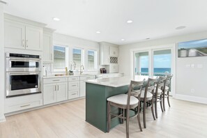 A modern kitchen with built-in appliances, a large island with bar stools, and a view of the ocean through the windows. - Oceanfront | 10 Bedrooms | Game Room | Pool | Theater (Emerald Isle)