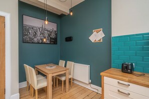 Ein kleiner Essbereich mit einem Holztisch und -stühlen, einem Stadtbild als Wandkunst, einem weißen Heizkörper und einer türkis-blauen Ziegelwand.