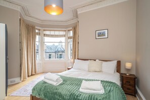 Ein Schlafzimmer mit einem großen Bett, einem Nachttisch, einer Lampe, einem Fenster mit Vorhängen und einem gerahmten Bild an der Wand.