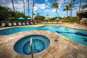 Ein Pool mit Liegestühlen und Sonnenschirmen, umgeben von Palmen und blauem Himmel.