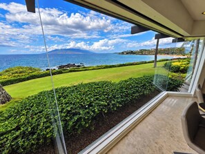 A view from a room with a large window overlooking a well-maintained lawn, a body of water, and distant mountains. - Stunning Ocean view -Wailea Point 2bd/2b Condo (Wailea)