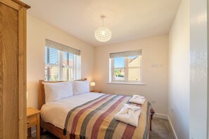 A bedroom with a wooden bed, a patterned blanket, a hanging light fixture, and windows with blinds. - Modern Home Near Blenheim Palace & The Cotswolds - Pass the Keys (Long Hanborough)