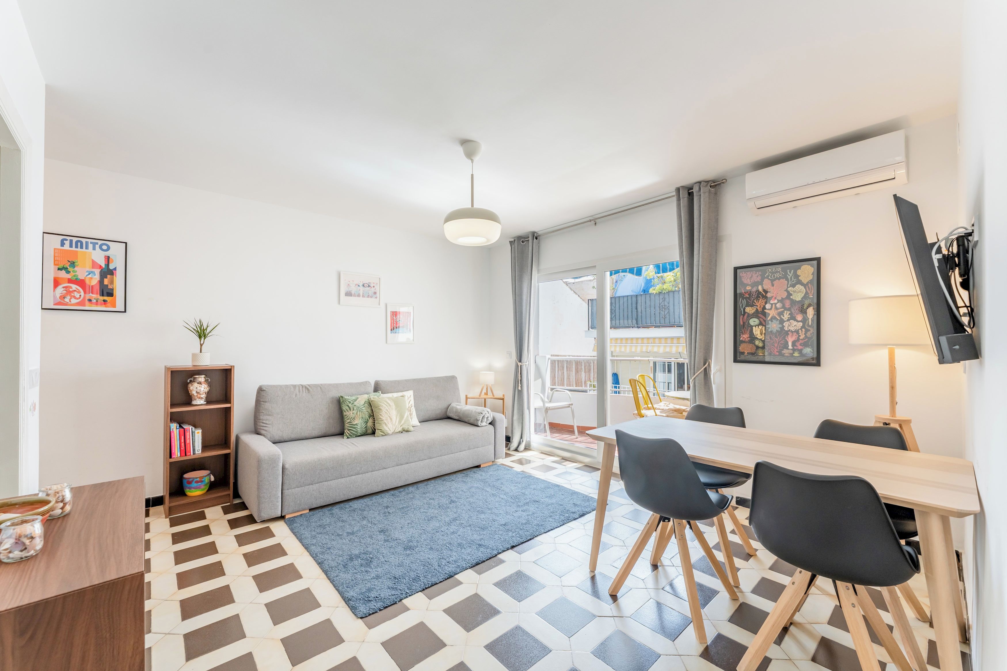 A modern living room with a grey sofa, a dining table with chairs, a patterned rug, and a bookshelf.