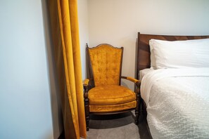 Habitació d'hotel amb llit de fusta, una cadira groga amb tapisseria i una cortina blanca.