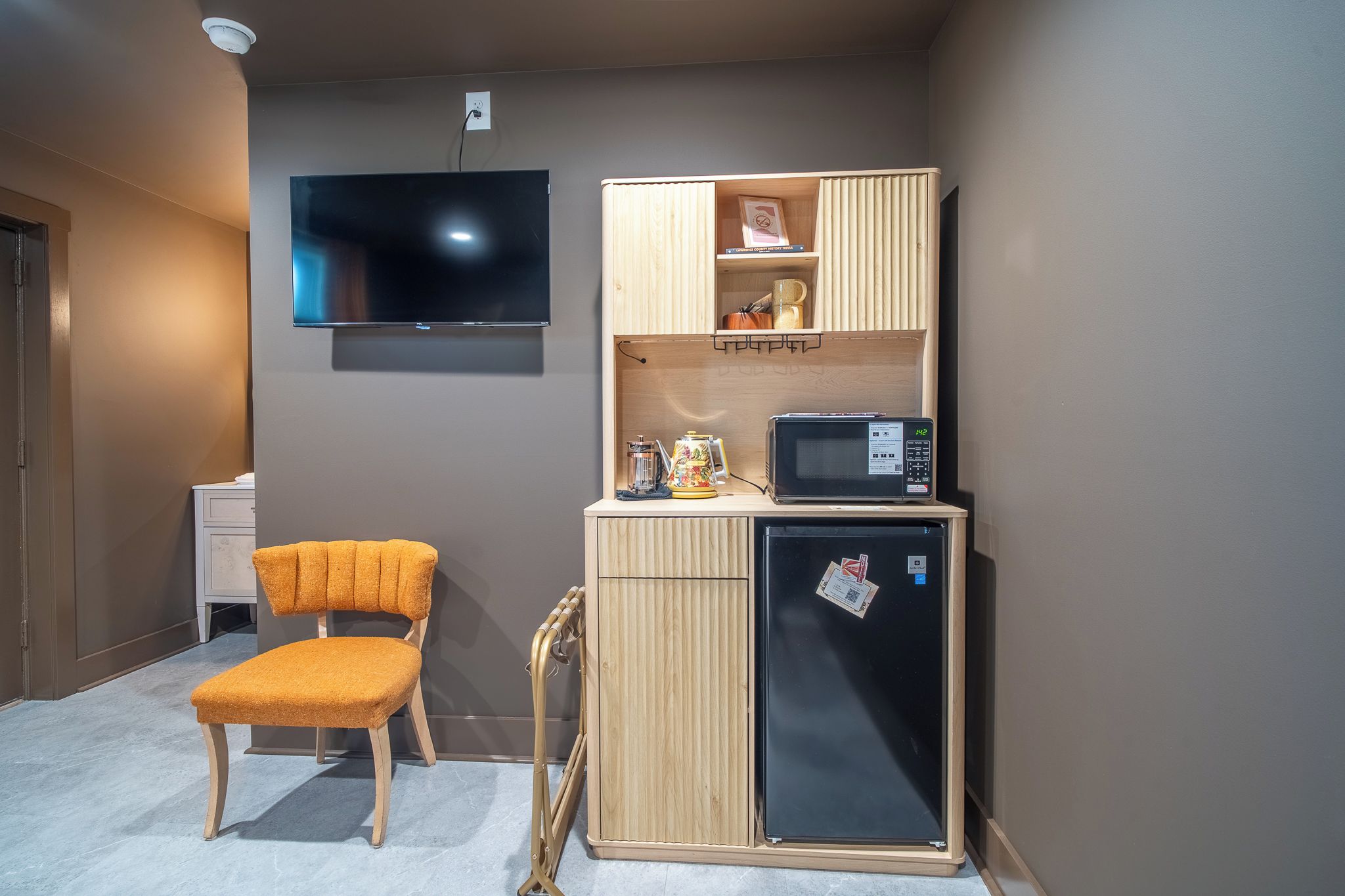 A compact kitchenette with a microwave, refrigerator, and a small shelf with items. A chair is placed in front of the kitchenette.