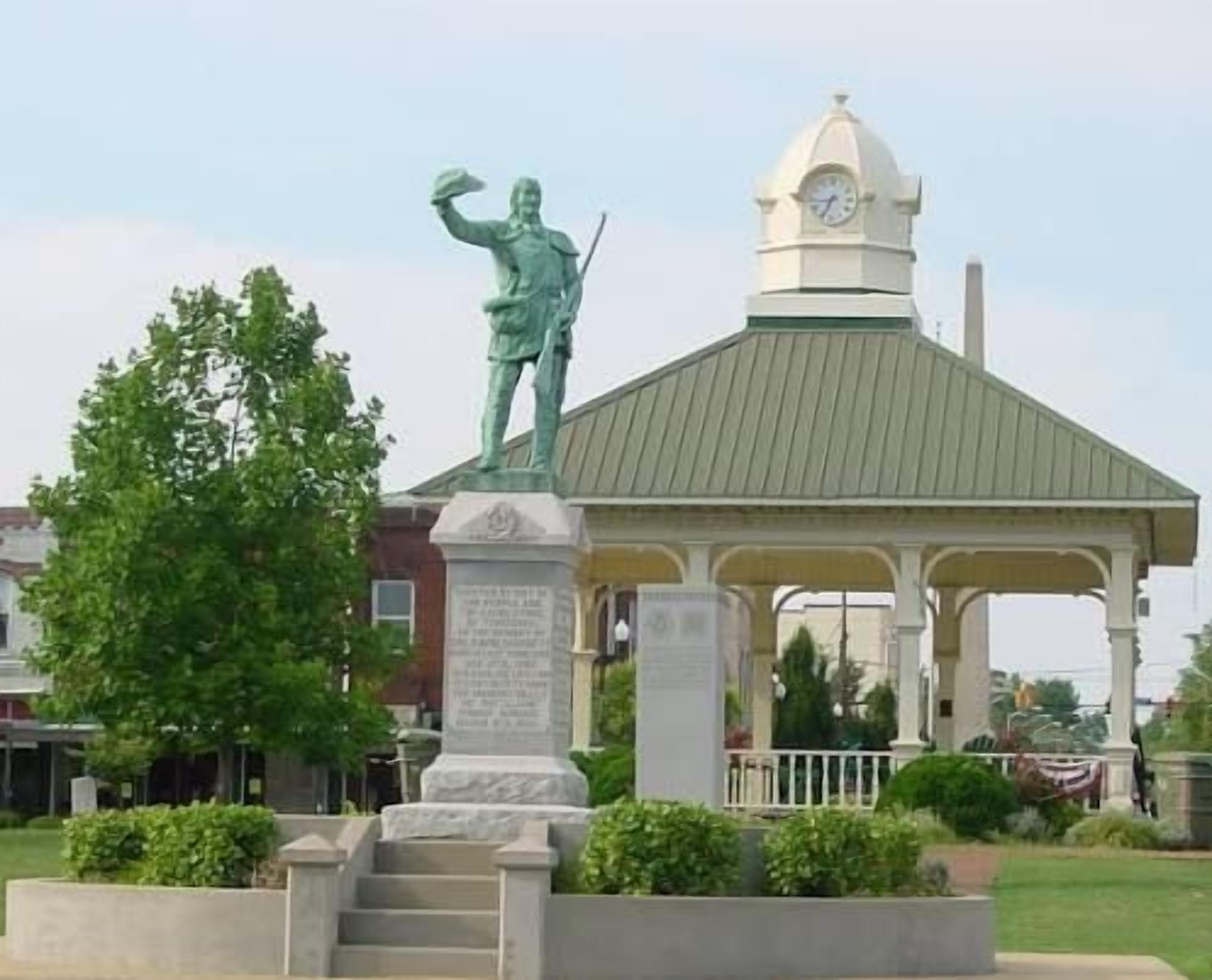A gazebo with a statue on top, surrounded by a small garden and a clock tower in the background.