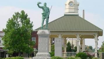 A gazebo with a statue on top, surrounded by a small garden and a clock tower in the background.