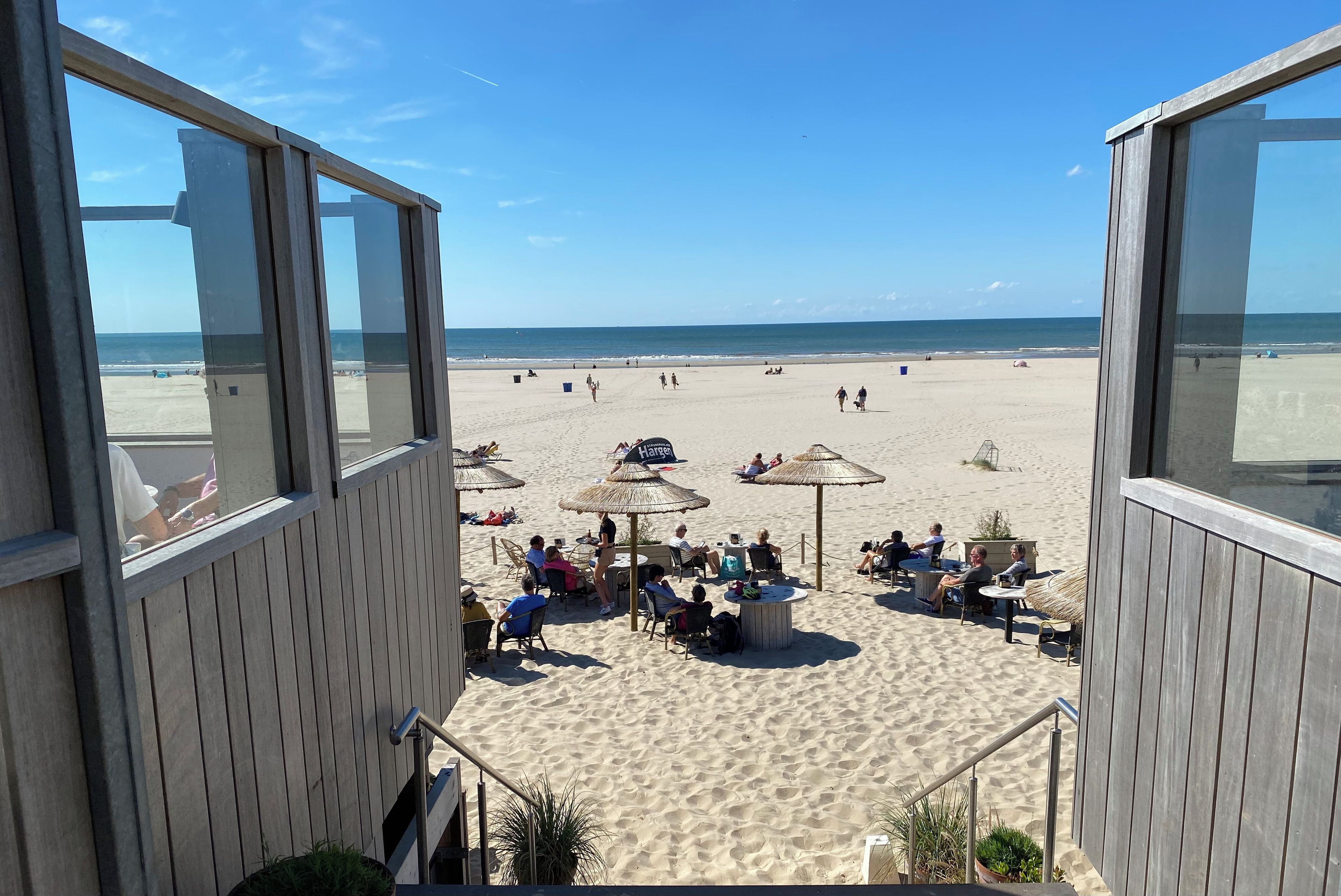 Une vue sur la plage depuis une terrasse en bois, avec des sièges d’extérieur et des parasols.