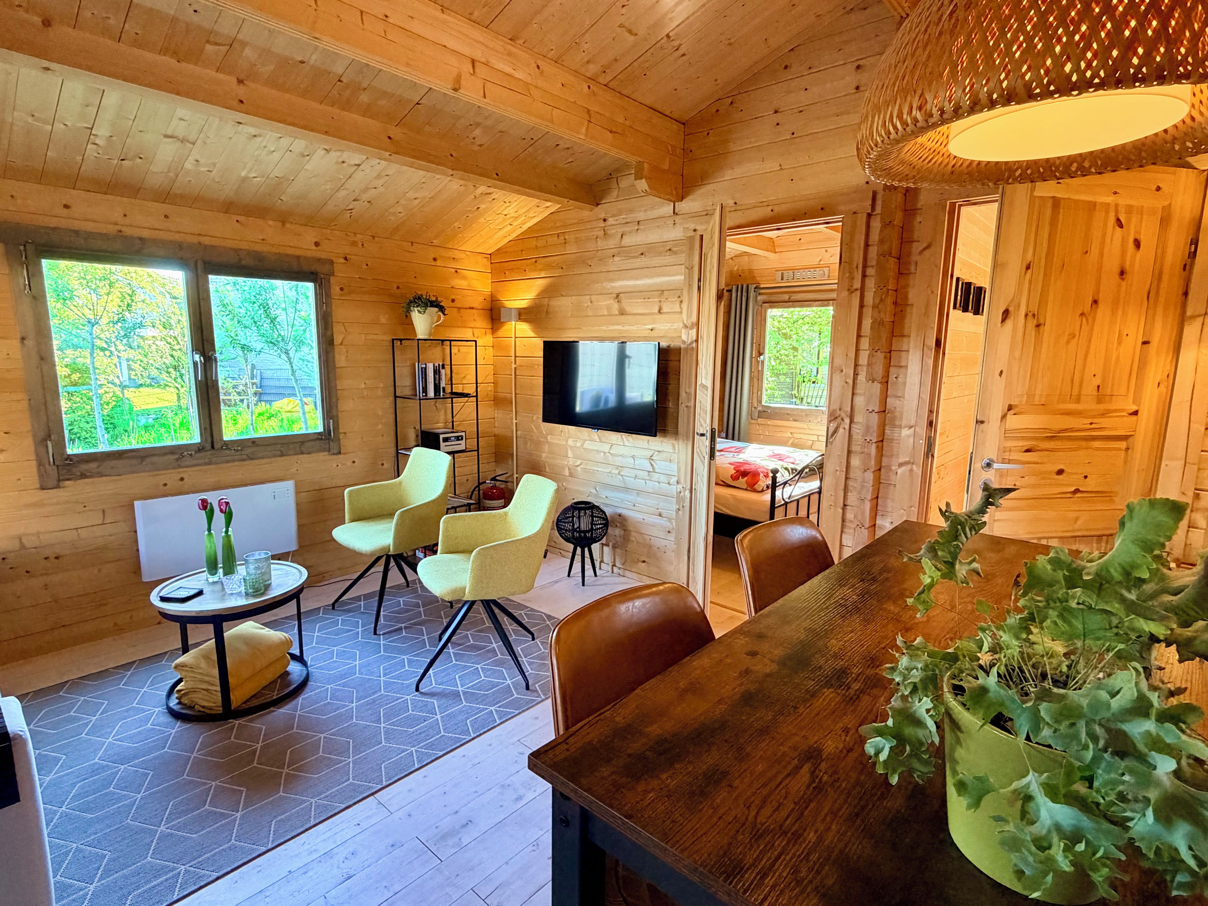 L’intérieur d’un chalet accueillant, avec des murs en bois, un coin repas, un téléviseur et une fenêtre donnant sur un paysage verdoyant.