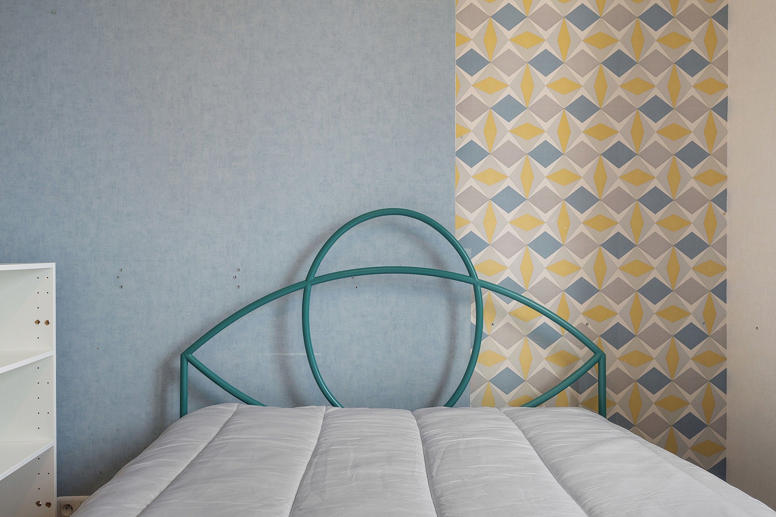A bed with a white mattress and a teal metal headboard against a wall with a geometric patterned wallpaper.