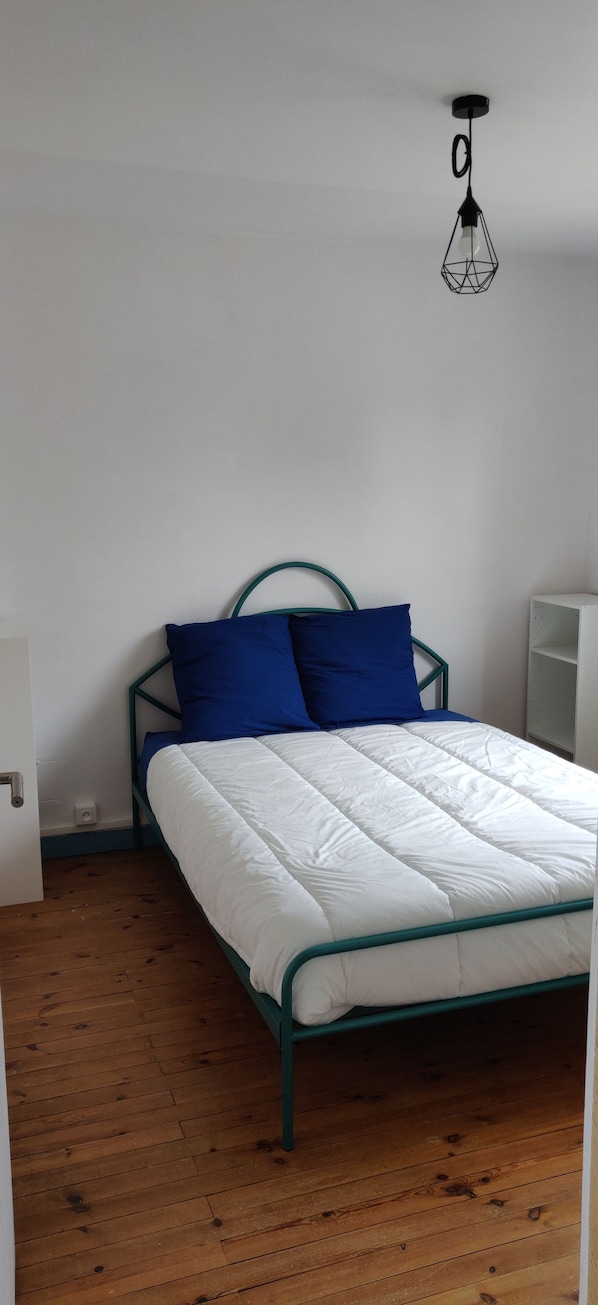Un lit simple avec un matelas blanc et un cadre de couleur turquoise, des oreillers bleus et un simple luminaire fixé au mur.