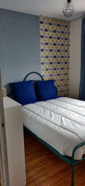 A single bed with a white mattress and teal metal frame, blue pillows, and a geometric patterned wall.