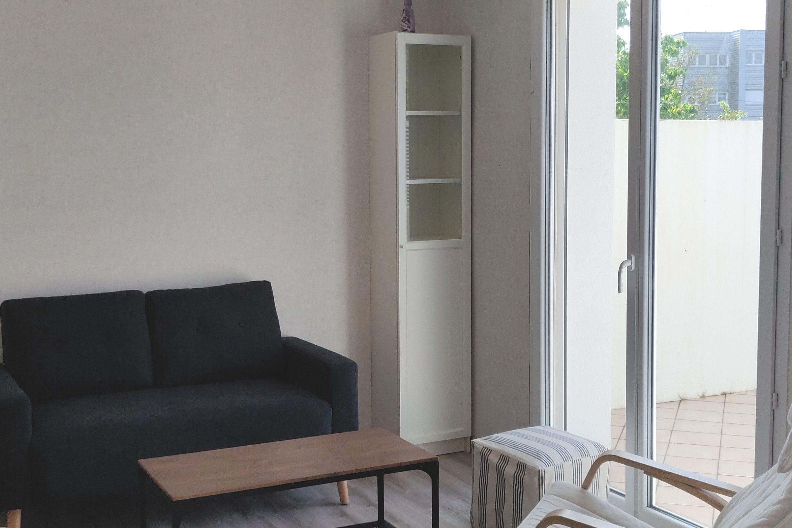 Ein Wohnzimmer mit einem dunklen Sofa, einem Holztisch, einer weißen Regaleinheit und Blick ins Freie durch eine Schiebetür.
