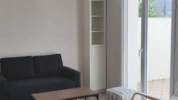 Ein Wohnzimmer mit einem dunklen Sofa, einem Holztisch, einer weißen Regaleinheit und Blick ins Freie durch eine Schiebetür.
