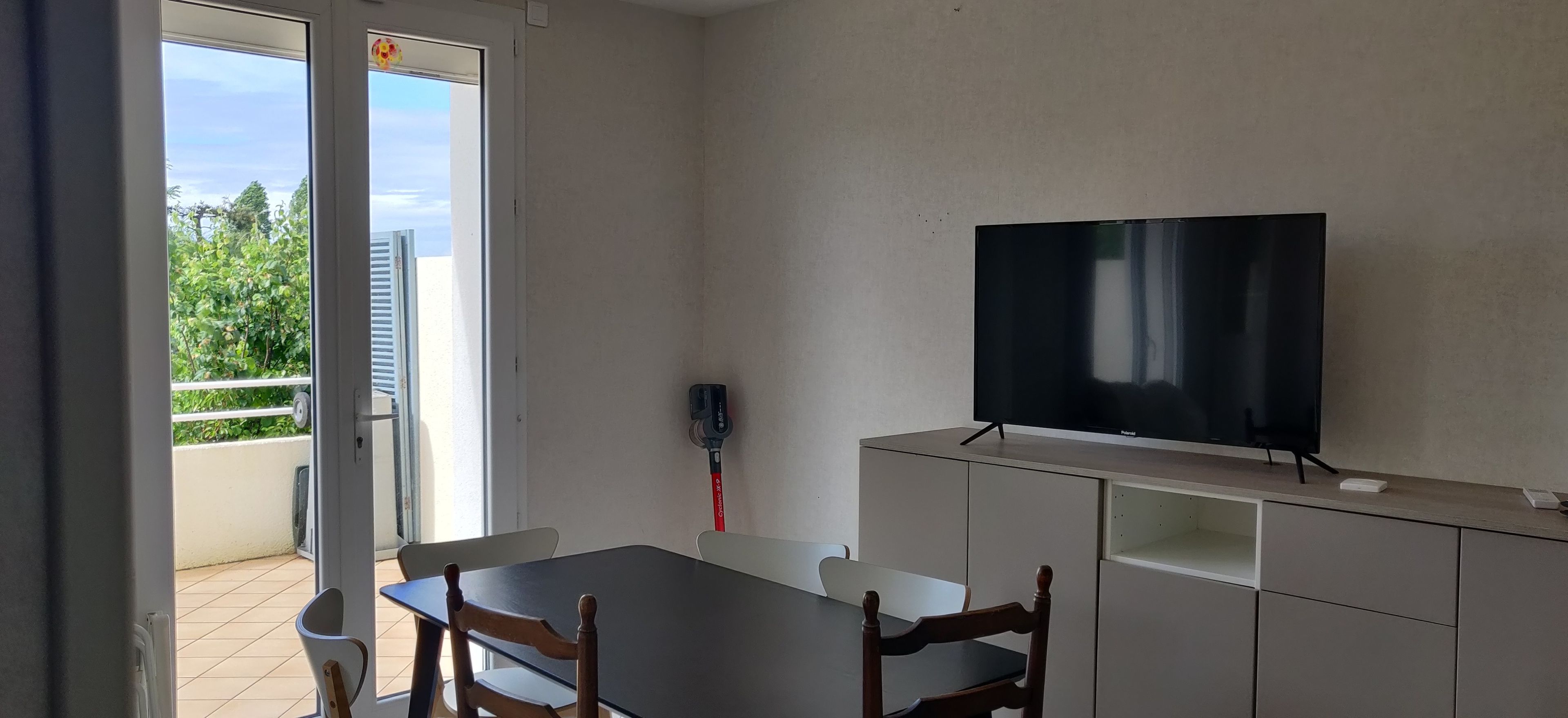 Ein Zimmer mit einem Fernseher auf einem Schrank, einem Esstisch mit Stühlen und einer Glasdoppeltür, die zu einem Außenbereich führt.