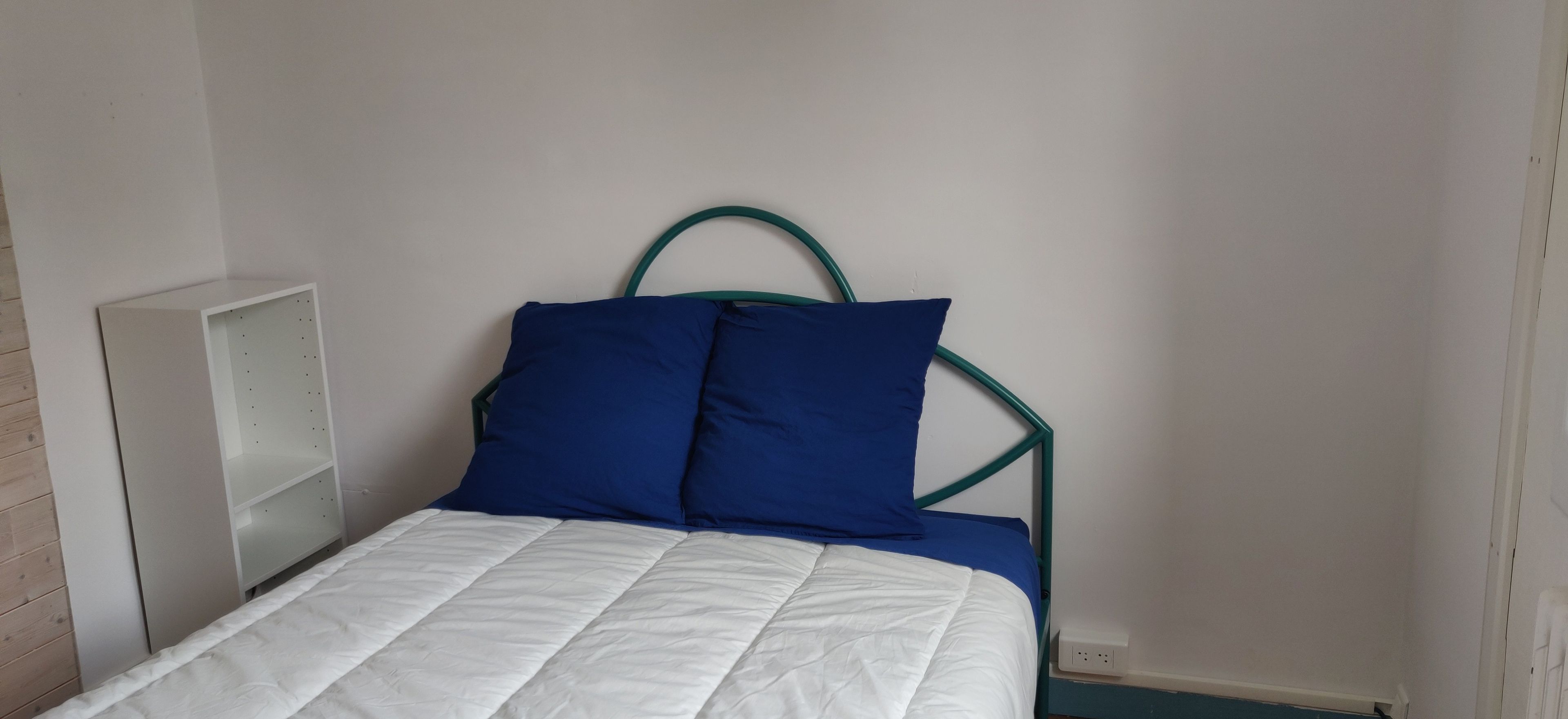 Ein Bett mit weißer Matratze und zwei blauen Kissen, ein Nachttisch in Weiß sowie ein grünes Metall-Kopfstück.