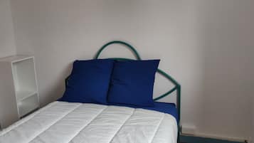 Ein Bett mit weißer Matratze und zwei blauen Kissen, ein Nachttisch in Weiß sowie ein grünes Metall-Kopfstück.