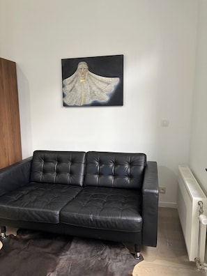 A black leather sofa with a tufted design, a black and white abstract painting on the wall, and a radiator on the floor. - Luxueuze en Cosy Appartement bij Atomium (Brussels)
