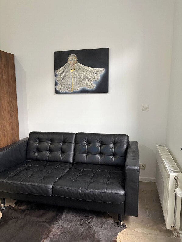 Um sofá de couro preto com design acolchoado, uma pintura abstrata em preto e branco na parede e um radiador no chão.