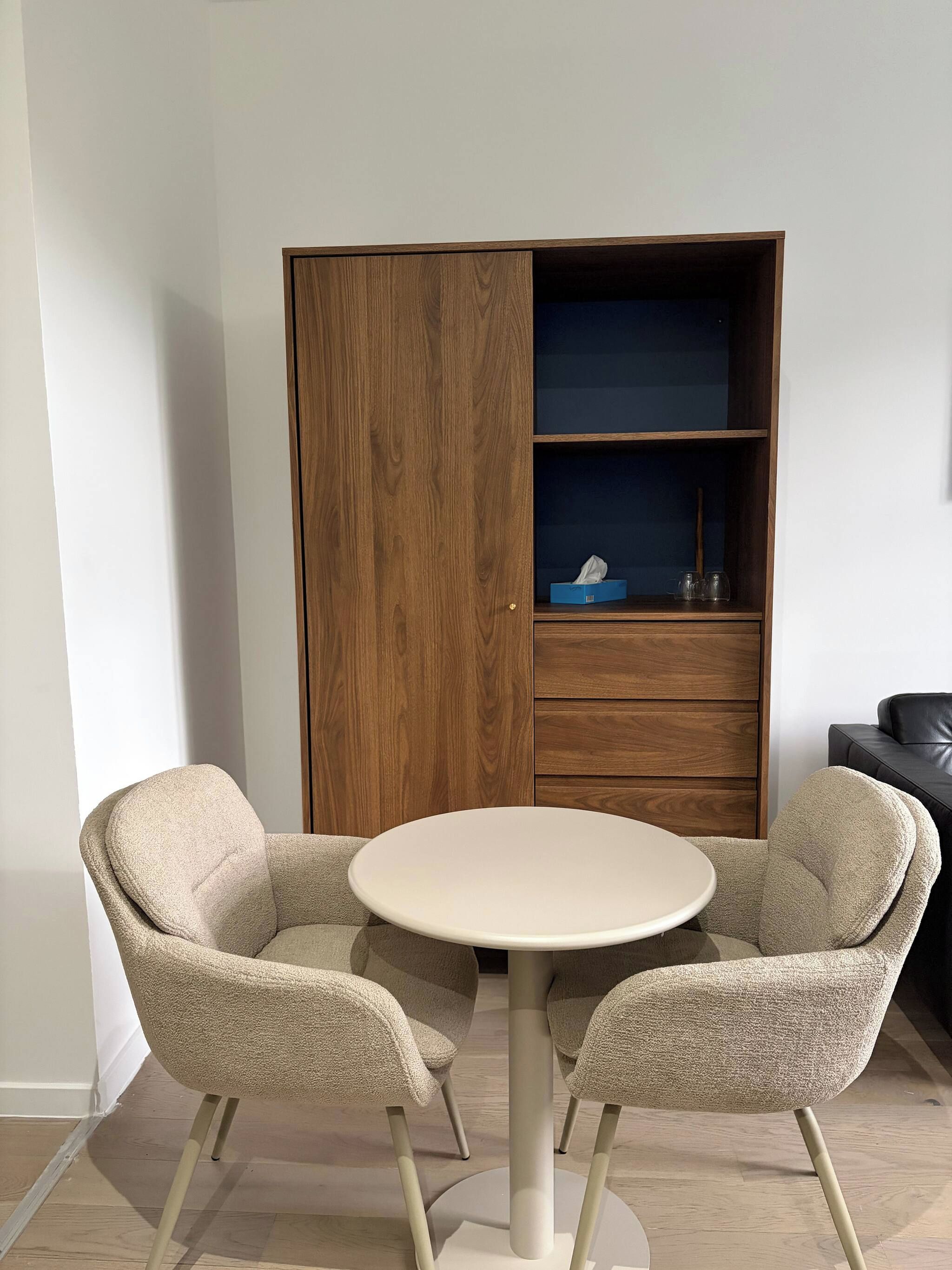 A room with a round table and two chairs, a wooden wardrobe, and a dark-colored sofa.