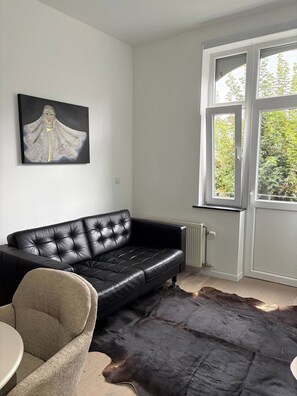Sala de estar com sofá de couro preto, poltrona bege, tapete preto e janela com vista para área verde.