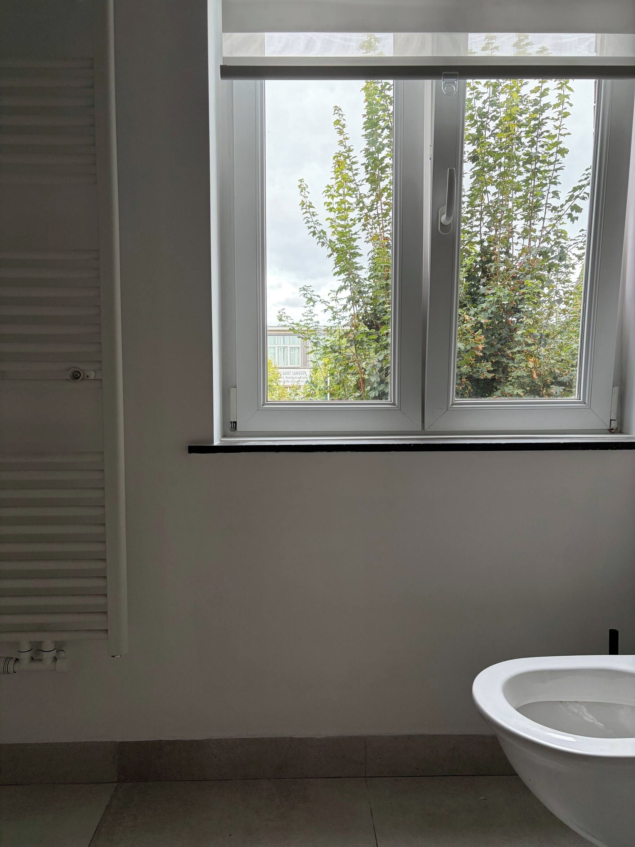 A bathroom with a toilet, a window with a view of trees and a building, and a radiator.