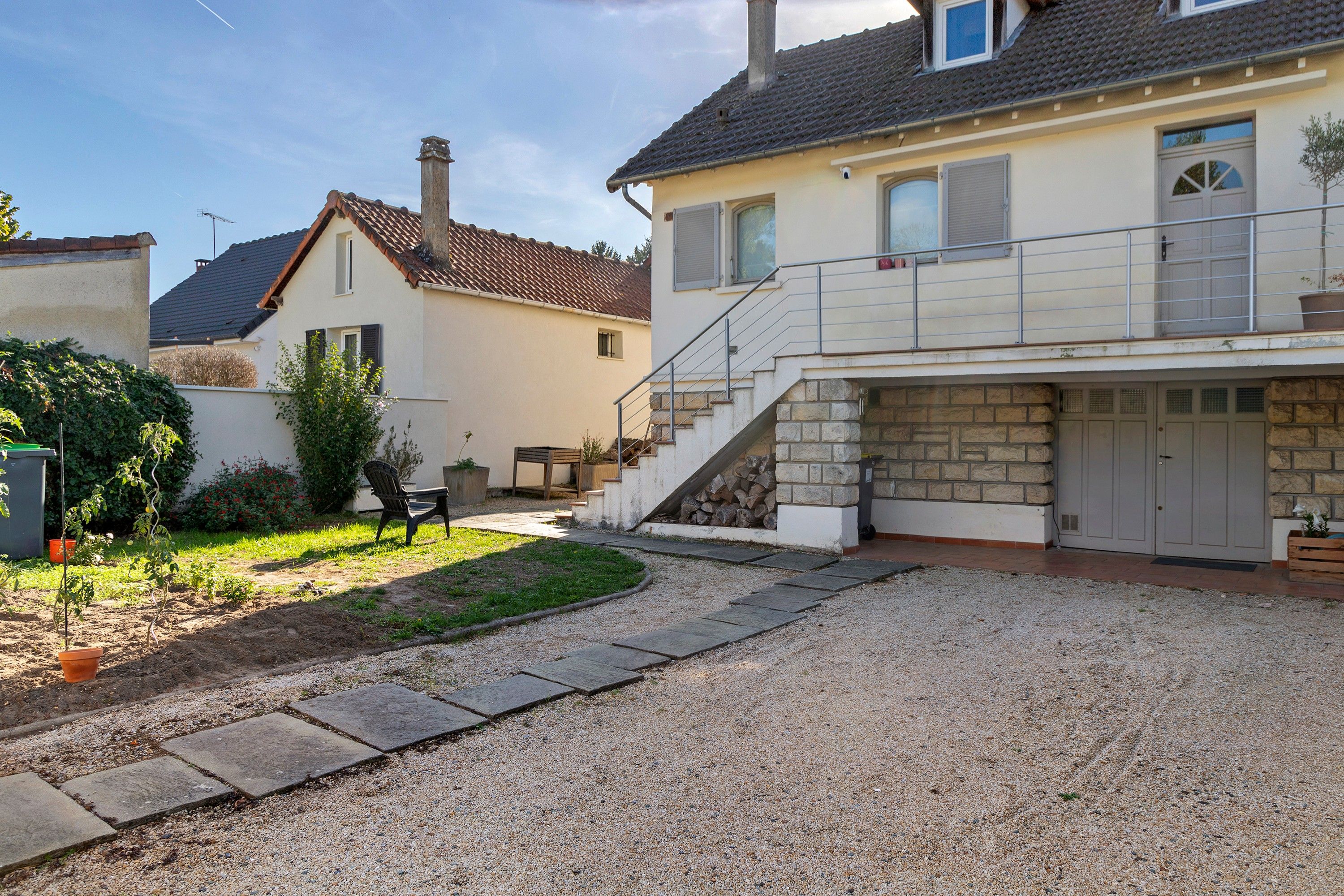 Il s'agit d'une propriété résidentielle dotée d'une allée en gravier, d'un escalier menant à un balcon situé au deuxième étage, et d'un jardin agrémenté d'une chaise et de plantes.